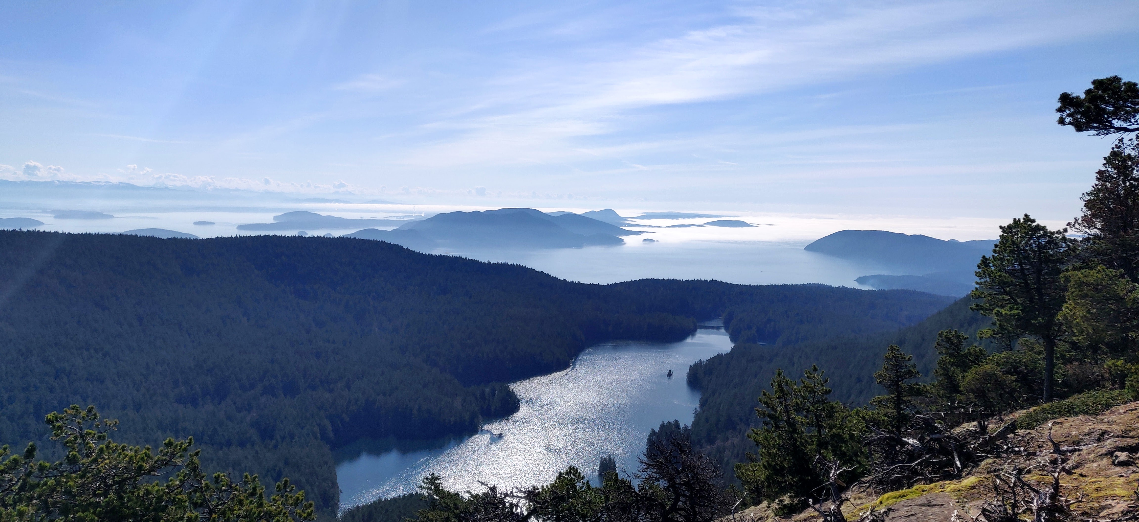 Aerial view from Mount Constitution: Mountain Lake in the foreground, surrounded by dense evergreen forest, with the San Juan Islands and blue water stretching into the distance beneath a clear sky.